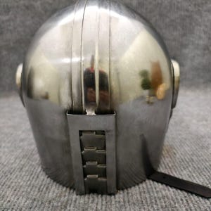 Puede incluir: Un casco plateado vintage con una parte superior redondeada y crestas verticales. El casco tiene piezas laterales y una secci&oacute;n trasera articulada con una correa de cuero. La superficie met&aacute;lica refleja el entorno.