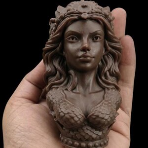 May include: A brown sculpture of a woman's bust, held in a hand. The bust features detailed hair, a crown, and a scaled bodice. The sculpture is a dark brown colour and is set against a black background.