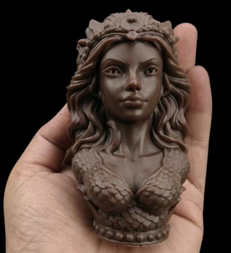 May include: A brown sculpture of a woman's bust, held in a hand. The bust features detailed hair, a crown, and a scaled bodice. The sculpture is a dark brown colour and is set against a black background.