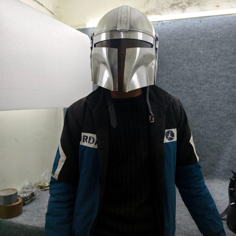 Puede incluir: Un casco mandaloriano plateado con una visera en forma de T. El casco se lleva sobre una chaqueta negra con detalles azules y la palabra "RD" visible en la manga. La chaqueta tambi&eacute;n tiene un logotipo con un jugador de baloncesto.