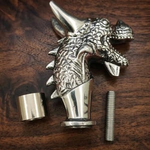 May include: A silver-colored metal dragon head handle with a textured scale pattern, featuring a smooth cylindrical collar and a separate threaded metal bolt. The dragon head has a prominent horn, open mouth, and sharp details across the surface.
