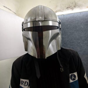 Puede incluir: Un casco mandaloriano plateado con una visera en forma de T. El casco tiene un acabado met&aacute;lico liso y una forma distintiva, con una cresta central y mejillas anguladas. La persona lleva una chaqueta negra con una franja azul y un logotipo.