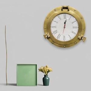 Könnte beinhalten: Eine messingfarbene Bullaugenuhr mit weißem Zifferblatt und schwarzen römischen Ziffern. Die Uhr hat einen roten Sekundenzeiger und ist an einer grauen Wand befestigt. Ein grünes quadratisches Tablett, eine kleine Vase mit Blumen und ein dünner brauner Stab befinden sich im Vordergrund.