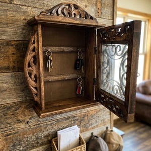 May include: A wall-mounted wooden key cabinet with a hinged door featuring a decorative carved frame and a glass panel. The interior contains two horizontal bars with metal hooks for hanging keys. Below the cabinet is a woven wicker basket.