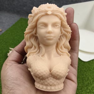 May include: A light beige bust of a mermaid with flowing hair and a crown. The mermaid's upper body is sculpted with scale-like details on the chest and shoulders. The sculpture is held in a hand against a green and white background.