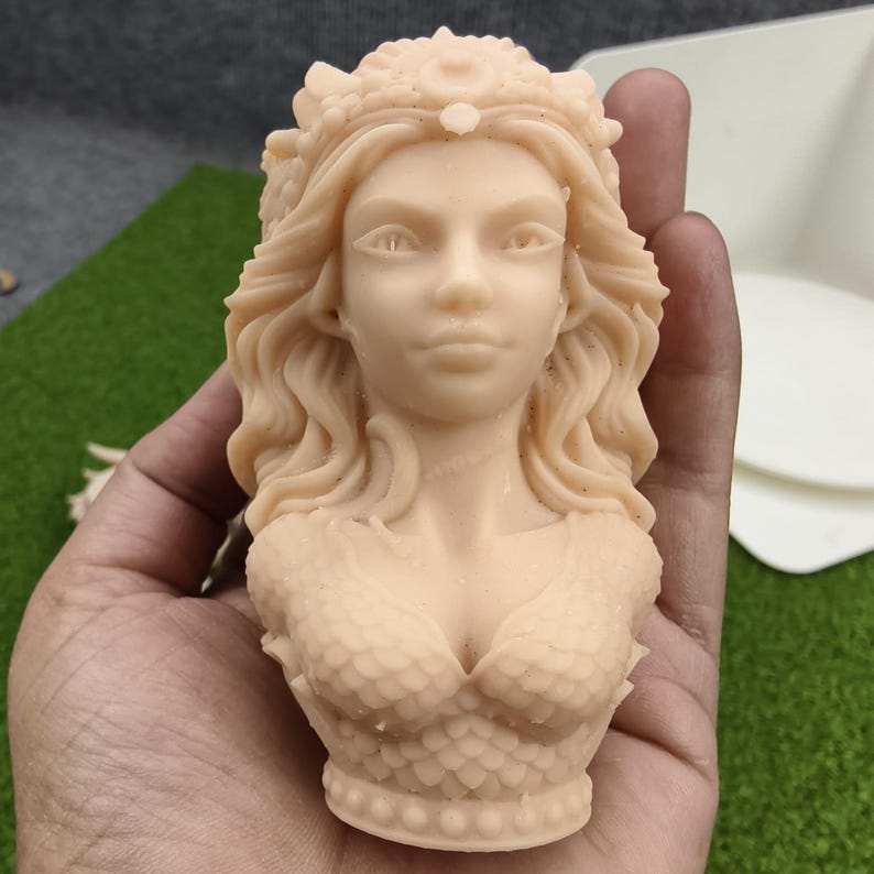 May include: A light beige bust of a mermaid with flowing hair and a crown. The mermaid's upper body is sculpted with scale-like details on the chest and shoulders. The sculpture is held in a hand against a green and white background.