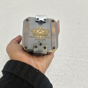 May include: A vintage metal compass with the text "KELVIN & HUGHES LONDON 1917" and a crown emblem. The compass has a silver and gold finish with brass accents. The compass is held in a hand.