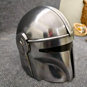 Puede incluir: Un casco plateado met&aacute;lico con visera y detalles en las orejas. El casco tiene una superficie lisa y reflectante y un dise&ntilde;o que recuerda a una est&eacute;tica espacial. El casco es probablemente un disfraz o un objeto de colecci&oacute;n.