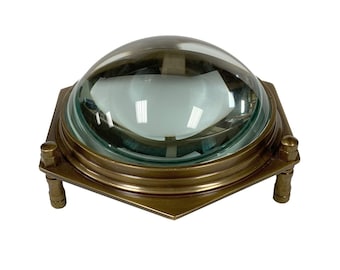 May include: A brass-toned, nautical-style dome light fixture with a clear, rounded glass lens. The fixture has a square base with rounded corners and is supported by four short, cylindrical legs.