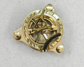 Puede incluir: Una brújula de reloj de sol dorada con números romanos y el texto "West London" grabado. La brújula presenta una aguja y una pieza negra curvada. El reloj de sol tiene tres perillas redondeadas.