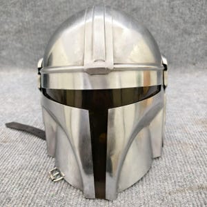 Puede incluir: Casco mandaloriano plateado con visera oscura. El casco tiene una distintiva visera en forma de T y una serie de crestas verticales en la parte superior. Una correa de cuero con hebilla est&aacute; sujeta al lateral. El casco es de metal.
