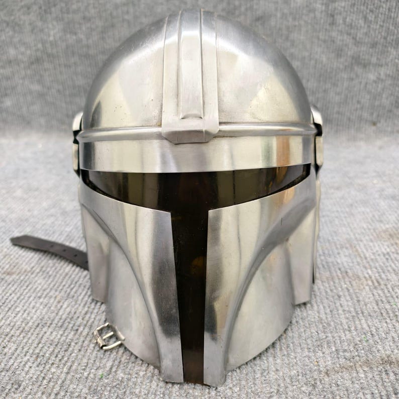 Puede incluir: Casco mandaloriano plateado con visera oscura. El casco tiene una distintiva visera en forma de T y una serie de crestas verticales en la parte superior. Una correa de cuero con hebilla est&aacute; sujeta al lateral. El casco es de metal.