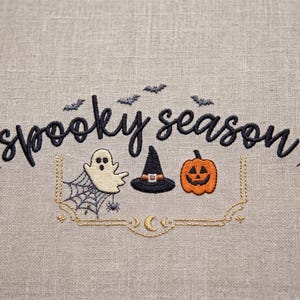 May include: An embroidery design on light beige fabric featuring the black text "spooky season" with small bats, a white ghost on a spider web, a black witch hat, an orange jack-o'-lantern, and purple leaf sprigs, framed by gold line art.