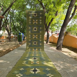 May include: A long, rectangular runner rug featuring a muted olive green base with a repeating geometric pattern in shades of blue, cream, and black. The rug has a flat-woven texture and is finished with short, dark fringe along the bottom edge.