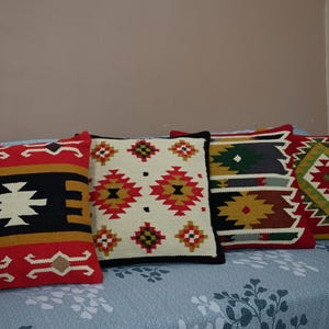May include: Four decorative throw pillows with Southwestern geometric patterns in red, black, white, yellow, and green. The pillows are arranged on a blue floral patterned sofa, offering a cozy and colorful accent for home decor.