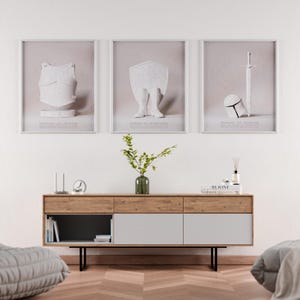 Könnte beinhalten: Drei gerahmte Kunstdrucke mit weißer Rüstung, einem Schild mit Beinen und einem Schwert mit Helm. Die Drucke befinden sich über einem Holzschrank mit grauem und weißem Design. Eine Vase mit Grün und dekorativen Gegenständen befindet sich auf dem Schrank.