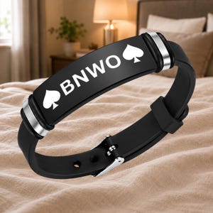 Könnte beinhalten: Ein schwarzes Silikonarmband mit einer zentralen rechteckigen Metallplatte in mattem Schwarz. Die Platte zeigt den weißen Text BNWO, flankiert von zwei weißen Pik-Symbolen. Das Armband ist mit silberfarbenen Metallakzenten und einer Schnalle versehen.