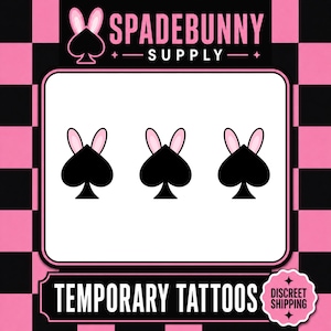 May include: Temporary tattoos with a black spade design and pink bunny ears. The image includes the text "SPADEBUNNY SUPPLY" and "TEMPORARY TATTOOS". The background has a pink and black chequered pattern.