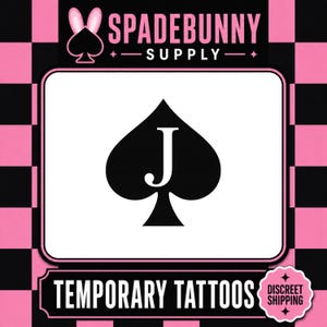 Op de afbeelding: Een witte rechthoek met een zwart schoppensymbool met de letter "J" in wit. De tekst "SPADEBUNNY SUPPLY" staat bovenaan en "TEMPORARY TATTOOS" onderaan. De achtergrond is een roze en zwart geblokt patroon.