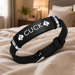May include: A black silicone bracelet featuring a central metal plate with the text CUCK and a spade symbol containing the Greek letter beta on each side. The bracelet includes silver-toned metal accents and a buckle closure for adjustment.