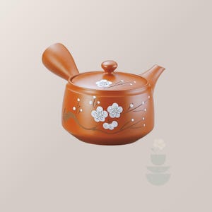 May include: A terracotta-colored teapot with a side handle and a lid. The body and lid feature white painted plum blossom designs with brown branches. The teapot has a short, curved spout and a rounded shape, showcasing a traditional Japanese style.
