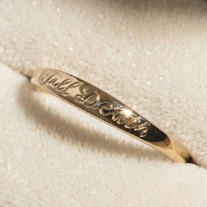 May include: A thin gold band ring featuring an engraved script inscription that reads Till Death. The metal has a polished finish and reflects light along the curved surface of the band, which is positioned diagonally across the frame.