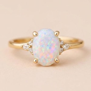 May include: A gold ring featuring a central oval-shaped iridescent opal stone held by four prongs. The band includes three small clear accent stones on each side of the center setting, creating a symmetrical design with a polished yellow gold finish.
