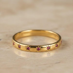 Könnte beinhalten: Ein goldener Ring mit einer Reihe von Sternenmustern, die jeweils einen kleinen, runden, roten Edelstein enthalten. Der Ring ist vor einem neutralen Hintergrund platziert, der seine polierte, reflektierende Oberfläche hervorhebt. Der Ring hat einen Durchmesser von etwa 1,8 cm.