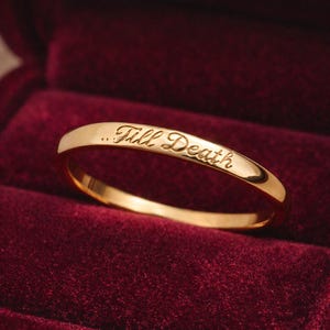 Puede incluir: Un anillo de oro con la inscripción "...Till Death" grabada en la superficie exterior. El anillo se muestra sobre una superficie de terciopelo rojo, destacando su acabado pulido y su diseño elegante. El anillo es un símbolo de compromiso.