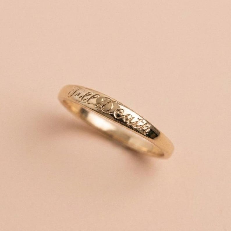May include: A polished gold band ring featuring the engraved cursive text Till Death along the outer surface. The ring has a smooth, rounded profile and a reflective metallic finish, showcasing the detailed script lettering etched into the metal.