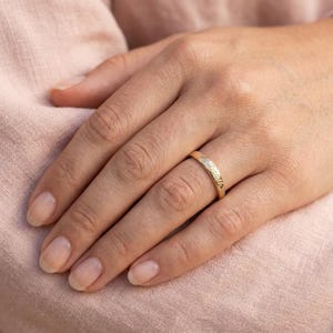 May include: A gold band ring featuring a smooth, polished surface with the word "Death" engraved in a cursive script font along the outer circumference of the metal band, which is worn on a finger against a soft, light pink fabric background.