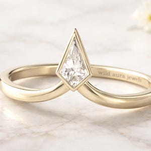 Puede incluir: Un anillo de oro con un diseño único. Presenta un diamante en forma de cometa engastado en un bisel. La banda lisa y pulida lleva grabada la inscripción "wild aura jewels". El anillo se muestra sobre una superficie de mármol.