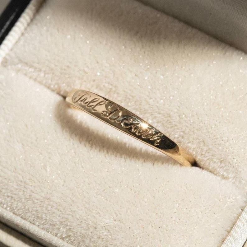 May include: A thin gold band ring featuring an engraved script inscription that reads Till Death. The metal has a polished finish and reflects light along the curved surface of the band, which is positioned diagonally across the frame.