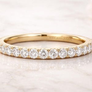 May include: A gold band ring with a continuous row of round, clear gemstones. The ring is set on a white surface. The text "wild aura jewels" is engraved on the inside of the ring.