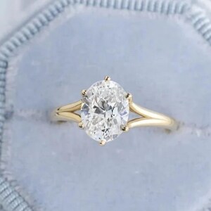 Puede incluir: Un anillo de compromiso de oro con un diamante de talla ovalada, engastado en una montura de seis garras. El anillo se muestra en un joyero de terciopelo azul claro. La banda tiene un diseño de vástago dividido.