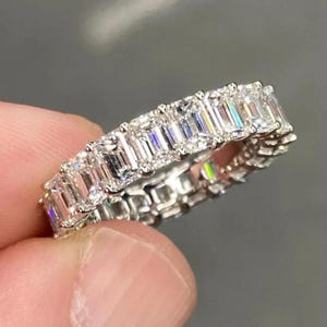 Può includere: Un anello eternity in argento con una fila continua di pietre preziose chiare taglio smeraldo. L'anello è tenuto tra le dita su uno sfondo sfocato, evidenziando la brillantezza delle pietre. Le pietre sono incastonate vicine, creando una fascia senza soluzione di continuità.