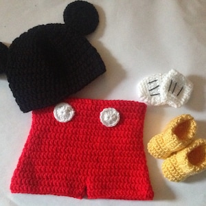Puede incluir: Un conjunto de bebé de ganchillo al estilo de un famoso personaje de dibujos animados. El conjunto incluye un gorro negro con orejas grandes, pantalones cortos rojos con botones blancos, guantes blancos y zapatos amarillos.