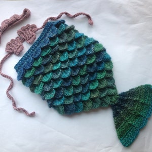 Puede incluir: Un disfraz de cola de sirena de ganchillo en tonos azules y verdes con una diadema a juego de ganchillo rosa.