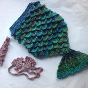 Puede incluir: Un disfraz de cola de sirena de ganchillo en tonos azules y verdes, con una diadema a juego y guantes sin dedos de color rosa polvoriento.