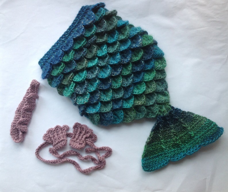 Puede incluir: Un disfraz de cola de sirena de ganchillo en tonos azules y verdes, con una diadema a juego y guantes sin dedos de color rosa polvoriento.
