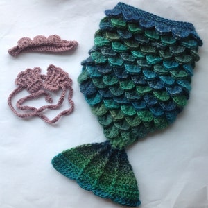 Puede incluir: Un disfraz de cola de sirena de ganchillo para un beb&eacute;, con diadema y pu&ntilde;os a juego. La cola est&aacute; hecha de hilo azul y verde, y la diadema y los pu&ntilde;os son de un rosa suave.