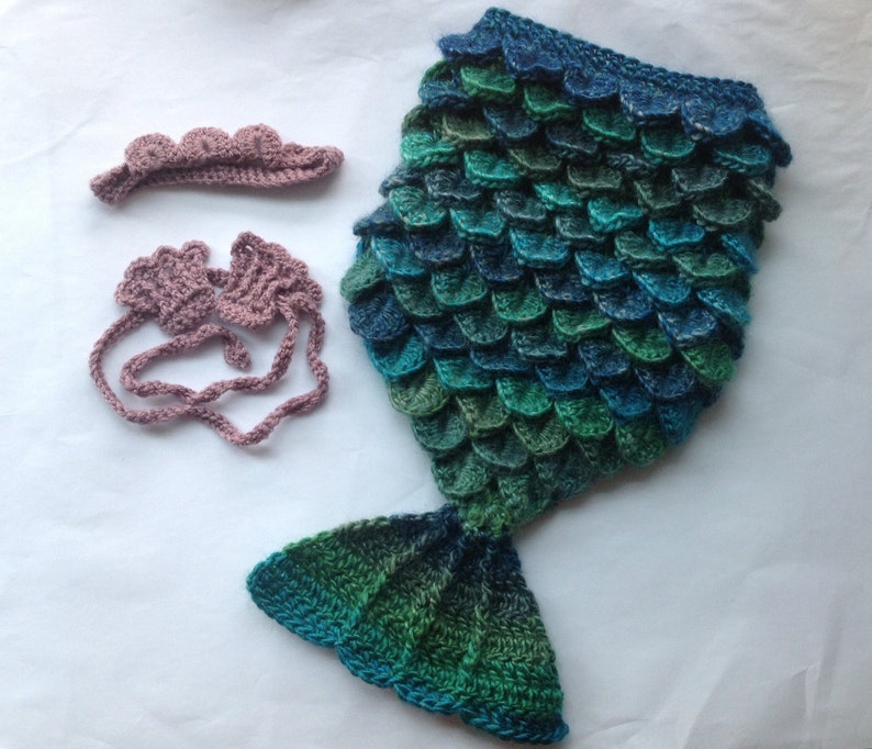 Puede incluir: Un disfraz de cola de sirena de ganchillo para un beb&eacute;, con diadema y pu&ntilde;os a juego. La cola est&aacute; hecha de hilo azul y verde, y la diadema y los pu&ntilde;os son de un rosa suave.