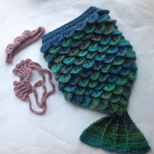Puede incluir: Un disfraz de cola de sirena de ganchillo en tonos azules y verdes con una corona y un collar a juego de ganchillo rosa.
