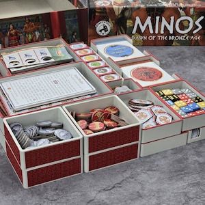 Könnte beinhalten: Ein Brettspiel-Organizer mit mehreren Fächern, die mit Spielkomponenten gefüllt sind. Der Organizer ist weiß mit roten Verzierungen und enthält Karten, Spielsteine, Würfel und Spielteile. Die Spielbox mit dem Titel "Minos: Dawn of the Bronze Age" ist im Hintergrund zu sehen.
