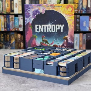 Könnte beinhalten: Ein blauer Brettspiel-Organizer aus Kunststoff mit mehreren Fächern, die verschiedene Spielkomponenten wie Kartenstapel und kleine Spielsteine enthalten. Hinter dem Organizer steht die Entropy-Spielschachtel mit farbenfrohen Illustrationen.