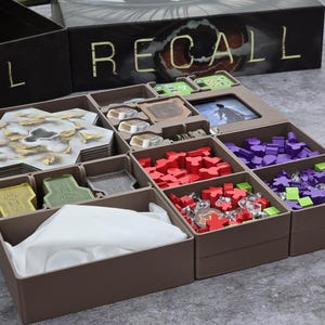 Könnte beinhalten: Ein brauner Spiel-Organizer mit mehreren Fächern, die mit Spielsteinen gefüllt sind. Die Fächer enthalten sechseckige Spielsteine, rote und lila Spielerfiguren und andere Spielkomponenten. Die Spielbox im Hintergrund trägt die Aufschrift "RECALL".