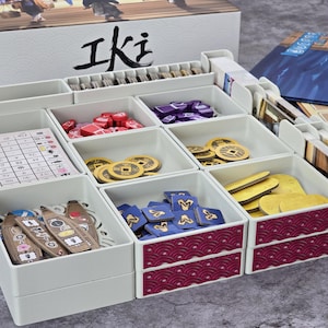 Könnte beinhalten: Ein weißer Brettspiel-Organizer mit mehreren Fächern, die mit Spielsteinen gefüllt sind. Die Fächer enthalten rote, lila und gelbe Spielsteine sowie Karten und Holzmarker. Die Spielbox ist im Hintergrund mit dem Wort "Iki" in Schwarz zu sehen.