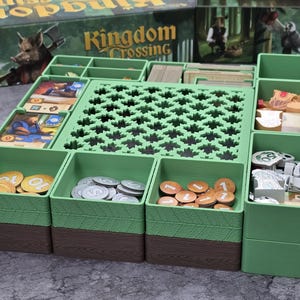 Könnte beinhalten: Ein grüner Brettspiel-Organizer mit mehreren Fächern. Der Organizer hat eine Holzstruktur auf der Unterseite und enthält Spielsteine, Karten und Token. Die Spielbox im Hintergrund trägt die Aufschrift "Kingdom Crossing".
