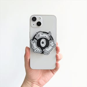 Könnte beinhalten: Ein weißes Smartphone mit einem schwarz-weißen Hunde-Handyhalter. Der Halter zeigt drei Zeichentrickhunde in einem Kreis um einen Pfotenabdruck. Der Text "JR VIP" ist sichtbar.