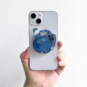 May include: A blue cat phone grip attached to a silver smartphone. The cat is depicted from the rear, with a white and blue striped tail. The phone has a dual-lens camera.